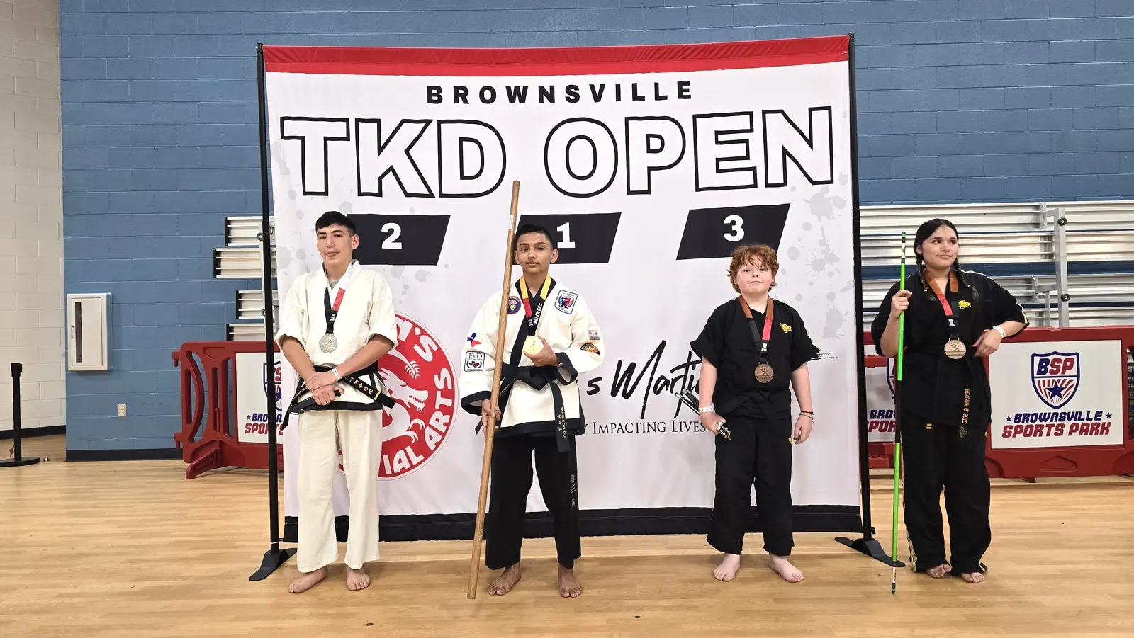 Adult and young taekwondo winners in white and black uniforms on the Brownsville TKD Open podium.