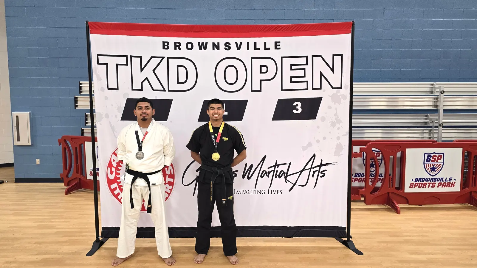 Two adult black belts with medals stand on the Brownsville TKD Open podium.