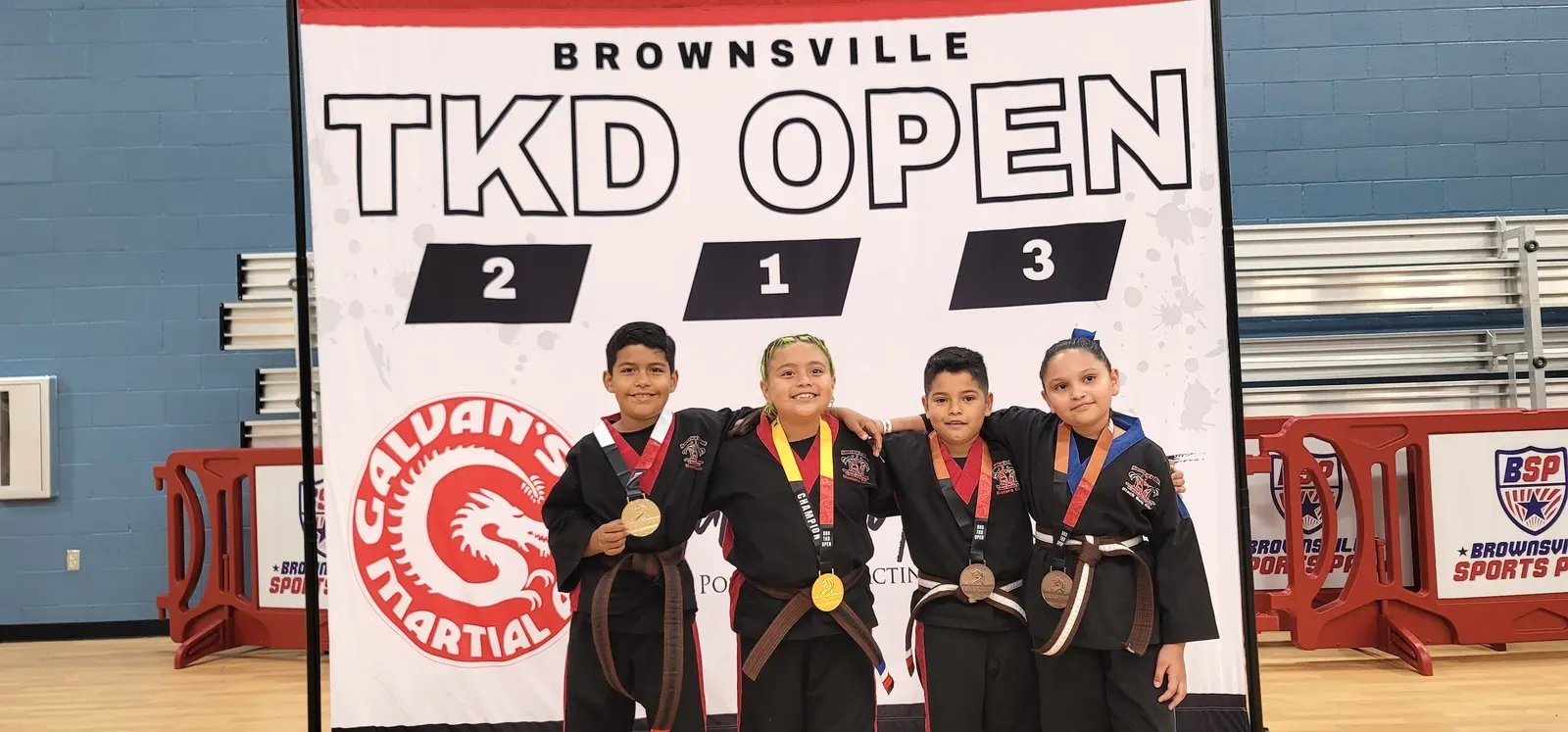 Four young taekwondo competitors in black uniforms wear their medals on the Brownsville TKD Open podium.
