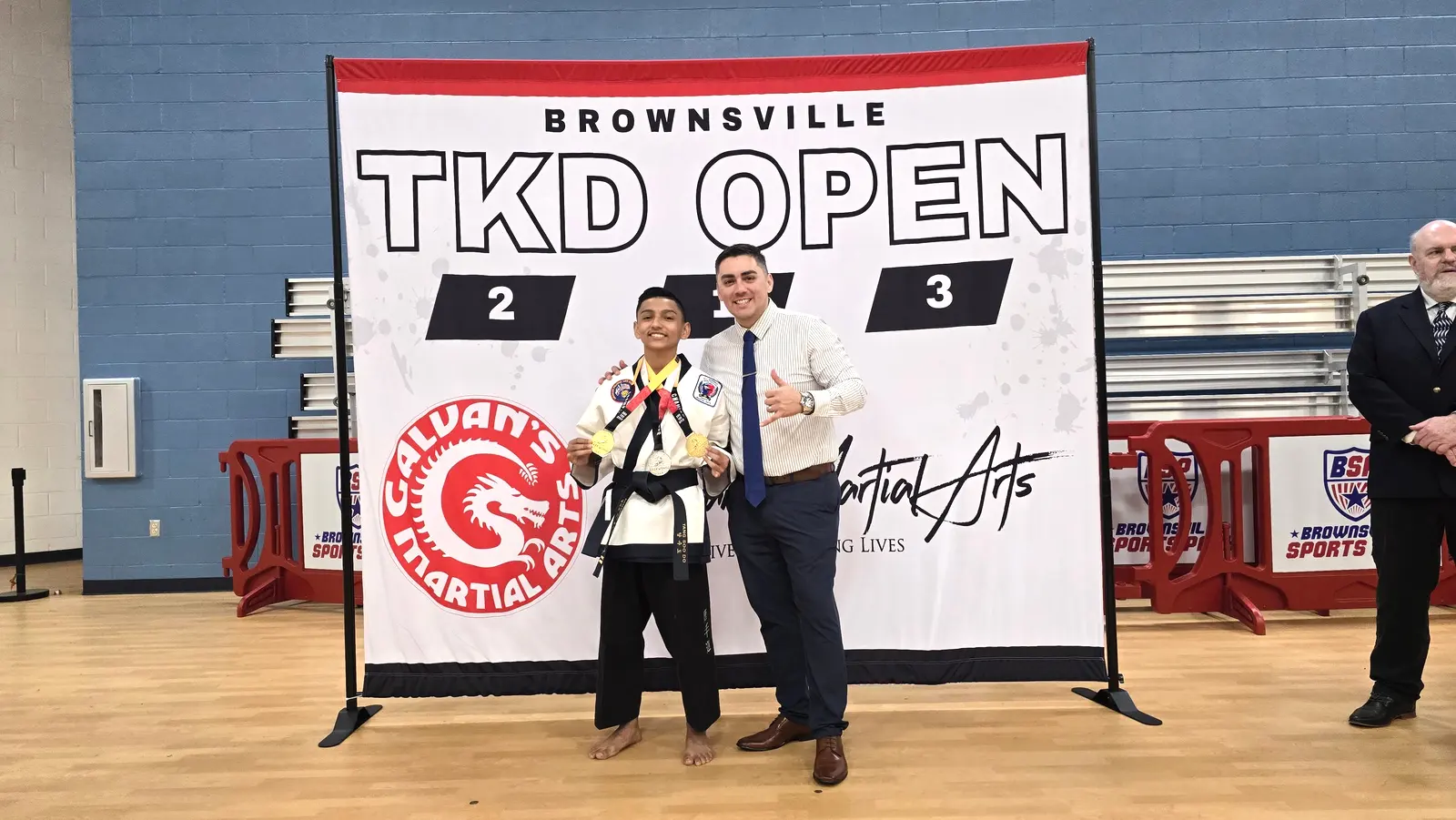 A young black-belt competitor with a medal stands beside his coach in front of the Brownsville TKD Open podium.