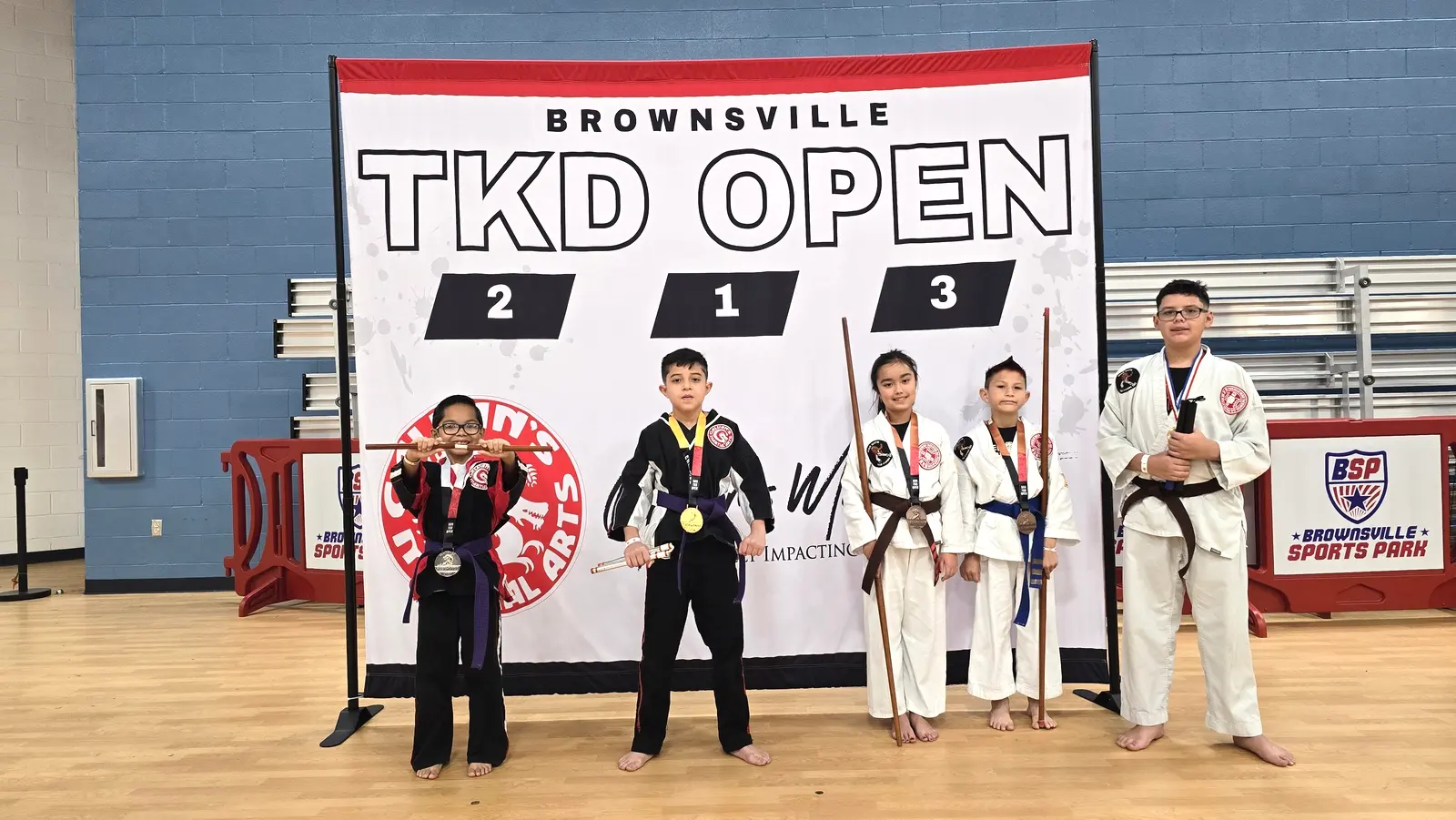 Young competitors holding weapons and wearing medals line up with their coaches on the Brownsville TKD Open podium.