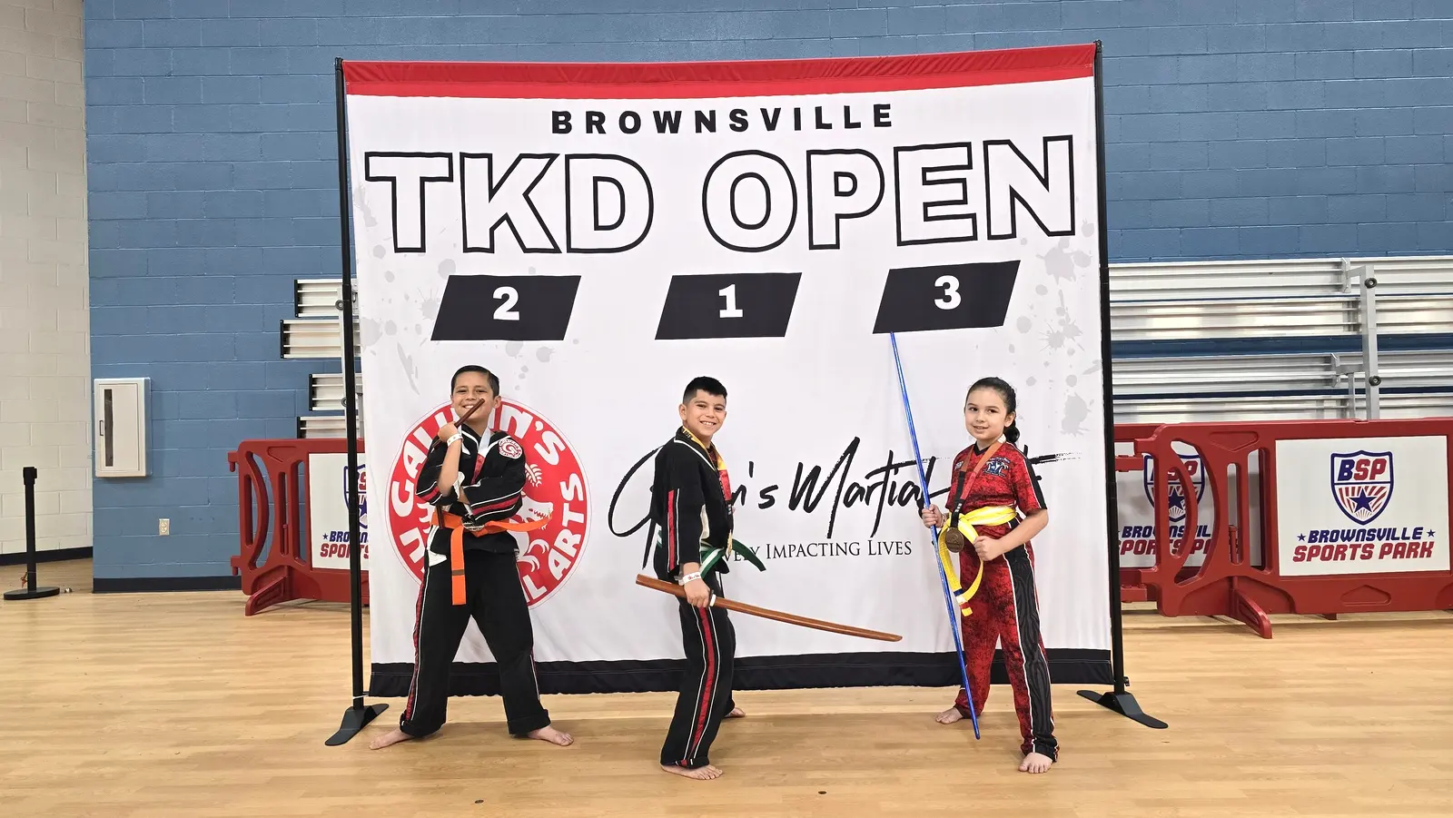 Two young taekwondo winners in red-trimmed uniforms stand with their instructors in front of the Brownsville TKD Open backdrop.